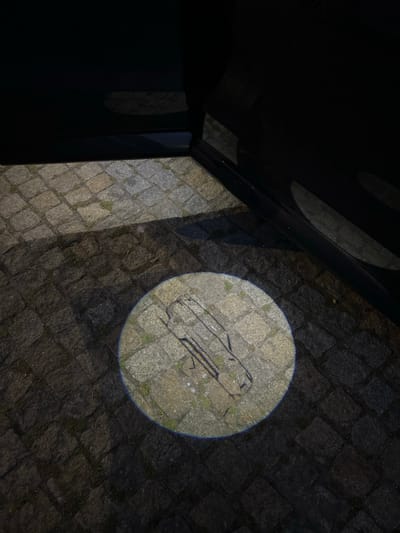 The image displays a low-light scene on a cobblestone surface in Caminha, Portugal. A dark vehicle's open door is visible in the upper and right portions of the frame. A circular light projection emanates from this open door, illuminating a section of the cobblestone ground below. Within this illuminated circle, a dark, stylized outline of a car, viewed in profile, is distinctly projected onto the stones. The cobblestones vary in tone and texture, with minor vegetation visible between them, particularly within the projection's radius. The surrounding environment is dim, with shadows cast from the vehicle and other unseen objects. No individuals are present in the scene.