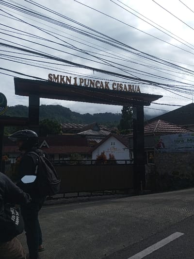 The image captures an outdoor scene featuring the entrance to a school, "SMKN 1 PUNCAK CISARUA," marked by a large archway sign. The sign, made of dark wood or metal, prominently displays the school's name in capital, light-colored letters. Above the archway, a dense network of black electrical or communication wires crisscrosses against an overcast, cloudy sky, indicating a dull or non-sunny day.
Behind the archway, a landscape of lush green hills and mountains is visible in the distance, partially obscured by mist or clouds, suggesting a mountainous region. Closer to the foreground, several buildings with distinctive reddish-brown tiled roofs are nestled among trees. One white building stands out with some faded, lighter-colored artwork or designs on its wall.
In the immediate foreground, a paved road stretches across the bottom of the frame, showing some textured patterns. On the left side of the road, two individuals are present. One person, seen from the waist up, is wearing a black helmet, a black jacket, and a black backpack, and appears to be sitting on a motorcycle, holding handlebars. To their left, another person is partially visible, wearing a light brown top and black shorts with white text that reads "GOOD TIMES HAPPY DAY" on the leg.
On the right side of the image, a low stone wall is visible, on which a banner is attached. The banner features an image of a man in traditional attire and text that reads "ISRA MI'RAJ NABI MUHAMMAD SAW," indicating a religious celebration. The overall setting suggests a school or institutional building located in a scenic, hilly, or mountainous area, likely in Indonesia given the text and architectural styles.