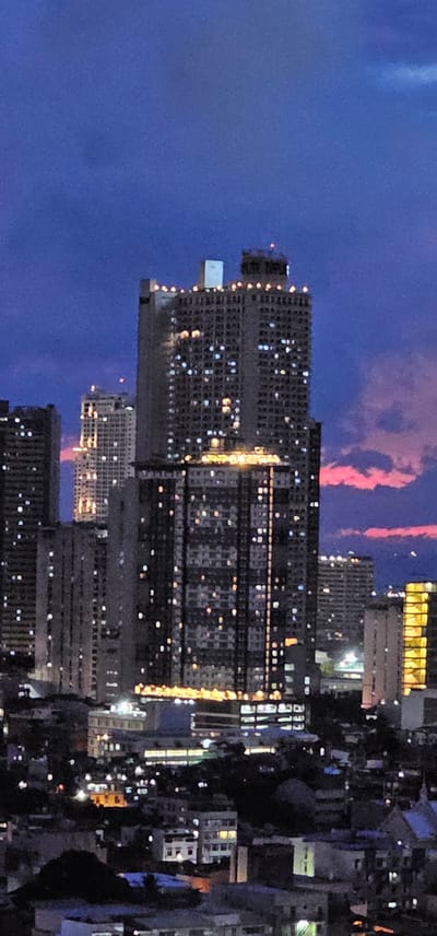 This media file captures an urban cityscape at dusk, with the sky transitioning from a deep blue to hues of pink and orange, suggesting a sunset or sunrise. The primary subjects are several tall buildings, some of which are illuminated with numerous lights, indicating that it is nighttime or twilight. The buildings are densely packed, typical of a metropolitan area. The scene is dominated by architecture, with few discernible objects or people.

The activity taking place is implied by the illuminated buildings, suggesting urban life continuing after dark. The time of day is clearly twilight or night, as evidenced by the dark sky and the lit windows of the buildings. The weather appears clear, with some wispy, pink-tinged clouds visible in the upper part of the sky. The location cues of dense urban development and specific architectural styles might point to a major city. However, without more specific visual information, pinpointing an exact location is difficult. There is no visible text in the image. The overall emotion conveyed by the image is one of urban vibrancy and the transition from day to night in a busy city.

The provided location context of Makati, Philippines, helps to interpret the scene as a view of a bustling business or residential district within that city. The dense arrangement of high-rise buildings is consistent with the urban landscape of Makati. The pinkish clouds in the sky are a natural phenomenon that can occur during twilight in various locations.