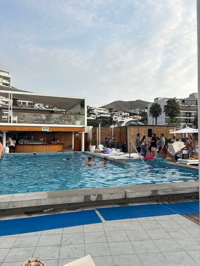The image shows an outdoor swimming pool at a seaside location in Ancón, Peru. The pool is lined with light blue tiles and appears to be a central gathering place, with people swimming and standing around it. Adjacent to the pool is a multi-story structure with a bar area, indicated by the presence of bar stools and a counter. "SALIDA" is visibly marked on the walls, potentially denoting an exit. People are gathered around the bar and pool area. The image features a mix of architectural elements, including white-framed buildings, bamboo paneling, and clear views of the sky and distant mountains.