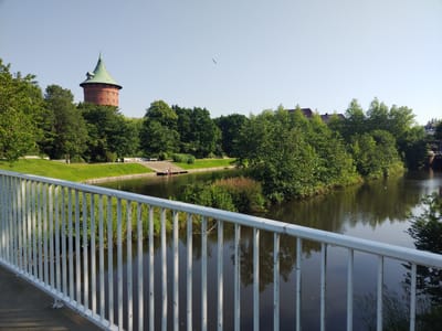 The image depicts a clear, sunny day scene at the Cuxhaven water tower in Germany. The water tower, constructed of red brick and topped with a green conical roof, is situated amidst lush green trees. In the foreground, a white metal railing runs horizontally across the frame, with vertical bars extending downwards. Beyond the railing, a dark river flows, reflecting the surrounding greenery and the sky. On the far bank of the river, more trees and some buildings with red roofs are visible. A few people are seated on the edge of a wooden dock on the riverbank. A bird is seen flying in the light blue sky. The environment suggests a tranquil park or riverside area within the city of Cuxhaven.