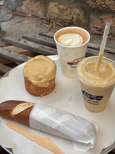 The image depicts an outdoor table setting at a cafe identified as "Coffee Wheels" (קפה גלגלים / Ha'Agala shel Kafe Galgalim), located in Kfar Vitkin, Israel. A round, silver-colored tray lined with white paper holds an assortment of breakfast or snack items. On the tray, from left to right, are: an elongated, dark brown pretzel-style bread roll partially wrapped in white paper, accompanied by a wooden disposable knife; a round pastry with a light brown base topped with a thick layer of creamy, light-colored frosting; a light brown paper cup containing a latte with a white heart-shaped latte art, adorned with graphics of coffee beans, croissants, and a stylized vintage van, along with Hebrew text "קפה גלגלים" (Coffee Wheels); and a clear plastic cup filled with a beige, textured smoothie-like drink, topped with a dome lid and a clear straw, also featuring the stylized van logo and Hebrew text "העגלה של קפה גלגלים" (The Cart of Coffee Wheels). The background consists of weathered wooden planks or slats, beyond which are blurred natural stone structures and dry vegetation. No individuals are visible in the scene.