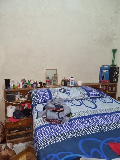 The image depicts an interior scene within a residence in Santa Cruz de la Sierra, Bolivia. The primary focus is a bed with a blue and white patterned comforter, which is partially covered by a pile of clothing. Beside the bed is a wooden shelving unit, densely packed with various items, including two Skyy Vodka boxes, a Heineken bottle, toiletries, personal care products, and miscellaneous household objects. A small mirror is attached to the wall near the shelf. The wall is painted in a light color. An individual appears partially visible in the lower right corner of the image.