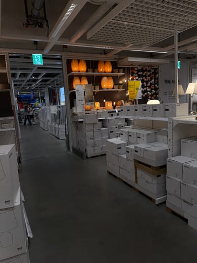 This is an indoor photograph of a large retail store, likely IKEA, located in Goyang-si, South Korea. The store features high ceilings with exposed industrial infrastructure and a grid-like ceiling panel system. Lighting fixtures are visible throughout, including long fluorescent tubes and decorative lamps.
In the foreground and midground, stacks of white cardboard boxes, many labeled with the IKEA logo and product names such as "ARSTID" and "BOJA," are arranged on pallets and shelves. These boxes appear to contain furniture or home goods. Several floor lamps with white shades are displayed on shelves, some in their packaging.
To the left, an aisle leads into the store, with more shelving and products visible in the distance. Two individuals, a male and a female, are walking away from the camera down this aisle. They are casually dressed, appearing to be shoppers.
On the right side of the image, a section of the store is dedicated to decorative items, with several woven pendant lamps in warm orange hues displayed on shelves. A yellow sign with the word "SALE" is also visible in this area. Further to the right, another floor lamp with a white shade is on display. A sign with Korean text, possibly indicating delivery services ("택배 서비스"), is also present.
An illuminated green exit sign with a running figure is mounted on a beam above the entrance to the store, indicating an emergency exit. The floor is a dark, polished concrete surface. The overall impression is of a well-organized retail environment showcasing a variety of home furnishings.