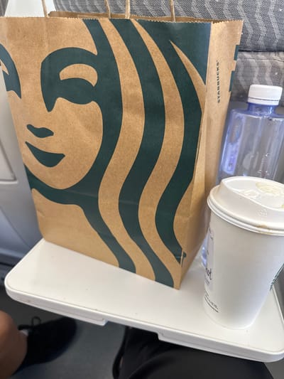 In the provided image, a Starbucks bag and a coffee cup are placed on a white tray table, likely in an airplane or a similar mode of transportation. The Starbucks bag features the iconic siren logo in green against a brown background. A white disposable coffee cup with a lid sits to the right of the bag, with some liquid residue on the lid. A partially visible blue water bottle is also present. The background shows a grey patterned fabric, possibly an airplane seat. The scene is captured from a close-up perspective, focusing on the items on the tray table. The image does not contain any specific visual cues that would indicate the location within the city of Dongguan, China.