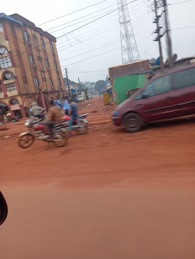 The image depicts a street scene in Imilike, Nigeria, characterized by its dusty red earth road. A multi-story building with brown and red striped facades, featuring numerous windows and air conditioning units, dominates the left side of the frame. Several motorcycles are visible on the road; one in the foreground is red and appears to be in motion, with two people on it. Another motorcycle is slightly behind it. A burgundy minivan is also in motion on the right side of the road. In the background, a large power line tower stands tall against a hazy sky, and various other buildings and trees can be seen. The overall impression is one of a busy, possibly commercial, area with a mix of residential and utility infrastructure.