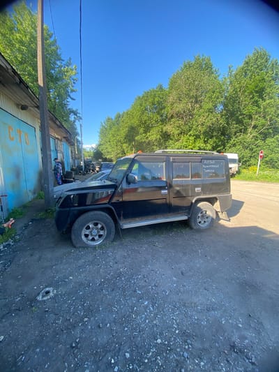 A black, older model SUV is parked on a gravel lot next to a light blue building with yellow lettering that reads "СТО". A person in a plaid shirt and colorful pants stands near the building, looking towards the SUV. The SUV has writing on its rear window that appears to be in Russian. Trees and a bright blue sky form the background. A red and white sign is visible in the distance. The overall scene depicts an outdoor area, possibly a service station or garage, in Kaykino, Russia.