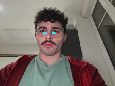 A person is upside down, wearing glasses, a light green shirt, and a red jacket. They have a mustache. Behind them is a window, a dark object, and a white wall with a square or rectangular fixture. The person's hair is curly and dark. Reflections are visible in the glasses.