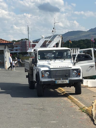 This daytime image captures a white Land Rover Defender parked sideways on an asphalt road in Saint-Jean-de-Luz, France. The classic Land Rover, with its driver's side door open, dominates the foreground. Its front grill clearly displays the "DEFENDER" branding, and the French license plate "DT-749-FL" prominently features the "64" department code, corresponding to Pyrénées-Atlantiques.

To the right of the vehicle, a yellow curb separates the road from a patch of ground where fishing nets are casually draped. Behind the Land Rover, a large white boat or ship is docked, featuring a prominent crane-like structure and various antennae, indicating a bustling maritime setting. Several flags, including a French tricolor and a yellow, red, and black flag, are visible on poles, likely associated with the boat or nearby structures.

In the mid-ground, a few casually dressed people are gathered near a building with a tiled roof, appearing to be observing the surroundings or simply relaxing. The background features rolling green mountains under a partly cloudy sky, characteristic of the region. The overall scene conveys a relaxed, sunny atmosphere typical of a coastal town in summer.