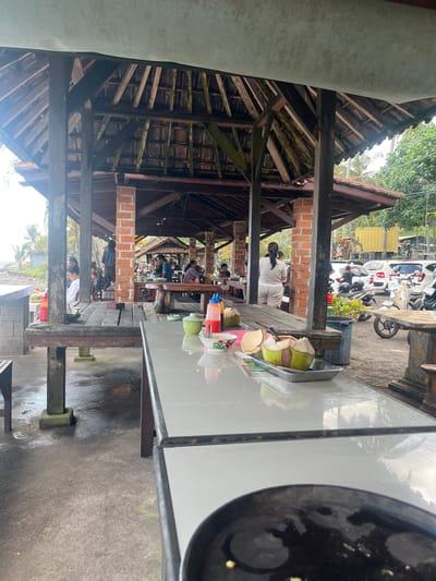 This image shows an outdoor seating area at what appears to be a casual eatery or restaurant, possibly in Pekutatan, Indonesia. The setting is characterized by a rustic, open-air structure with a wooden roof and supporting beams, suggesting a tropical environment. Several people are seated at tables, engaged in conversation or eating.
In the foreground, a glass-topped table is visible, with a tray holding a young coconut and some cut coconut pieces. The table surface reflects the sky and surrounding greenery, hinting at daylight hours. Beyond the table, the covered seating area stretches back, with more tables and patrons. The architecture features exposed wooden beams and terracotta brickwork.
In the background, several motorcycles and a white car are parked, indicating this might be a place accessible by road. Lush green foliage can be seen, further suggesting a warm, possibly humid climate. The overall atmosphere appears relaxed and informal. There is no discernible text in the image.