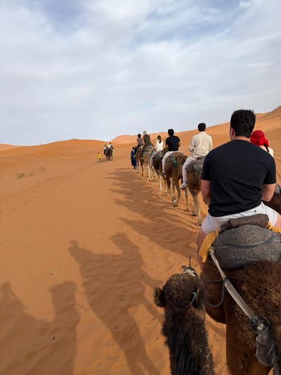 A line of people are riding camels through the desert on sand dunes. The sky is cloudy. The location is the city of Rissani, Morocco.