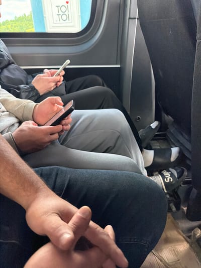 The image depicts the interior of a vehicle, possibly a bus or train, with passengers seated. The perspective is from within the vehicle, looking forward and slightly to the side. In the foreground, a person's arms and hands are clasped together, resting on dark blue denim jeans. To the left and slightly behind, another person's forearms and hands are visible, holding a smartphone. Further back, another individual's hands are visible holding a similar device. Another person's legs are also visible, wearing grey sweatpants and black and white socks with the word "Supreme" visible on one of them. Through the window, a blurred outdoor scene with greenery and a white structure with text is visible. The overall environment suggests a journey. The presence of the "Supreme" brand on the socks and the general context of travel do not specifically pinpoint the location to Jodłownik, Poland, or any other specific city.