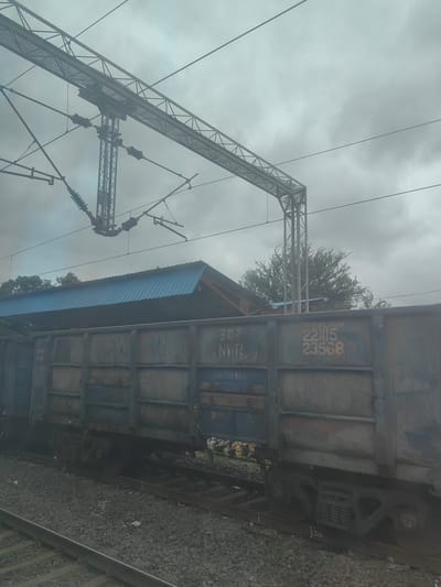A low-angle, slightly over-the-shoulder shot captures a train on a cloudy day in Shivpur, India. The foreground is dominated by a railway track with ballast. To the right, the side of a weathered, blue-grey freight train car is visible. Large yellow numbers "221115" and "23568" are stenciled on its side. Above the train car, a blue corrugated metal roof of a station platform stretches across the frame. Overhead, complex metal gantries and wires for an electric railway system crisscross the overcast sky, with some insulators visible. Lush green trees are visible in the background, behind the station roof and to the right. The overall scene depicts a typical railway environment under a heavy, grey sky, suggesting potential for rain.
