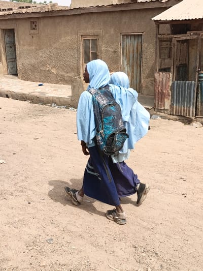 Two individuals are observed walking from left to right on an unpaved, dusty street in Zango, Nigeria. The lead individual wears a light blue head covering, a light blue long-sleeved garment, and a dark blue skirt, carrying a black and teal patterned backpack. Their footwear consists of worn athletic shoes. A second individual, also in light blue attire, is partially visible behind the first. In the background, to the left, stands a light brown, textured building with a light-colored wooden-framed door and a window. To the right, a structure constructed from corrugated metal and wood is present. Small fragments of debris are scattered on the ground near the base of the left building. Shadows cast by the individuals indicate strong overhead sunlight.