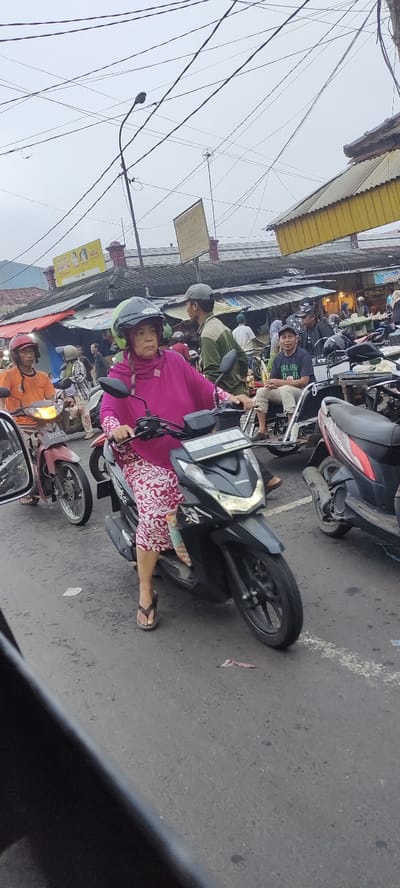 The image shows a street scene in Mojokerto, Indonesia (latitude: -7.468, longitude: 112.438). A woman wearing a pink hijab and a floral patterned dress is riding a black motorcycle. She is wearing a helmet and has a white scarf covering her hair. The motorcycle is stopped in traffic, and the woman is looking straight ahead. There are several other people in the background, including a man wearing a green shirt, a man wearing a black shirt, and a man wearing a blue shirt. There are several motorcycles parked in the background. The street is paved with asphalt and there are several power lines overhead. The sky is cloudy. The scene is busy and there is a lot of activity. The weather appears to be overcast. The scene appears to be in an urban setting. The woman appears to be in her 30s or 40s. The men in the background appear to be in their 20s or 30s. The image was taken from the perspective of a person in the street looking at the woman riding the motorcycle.