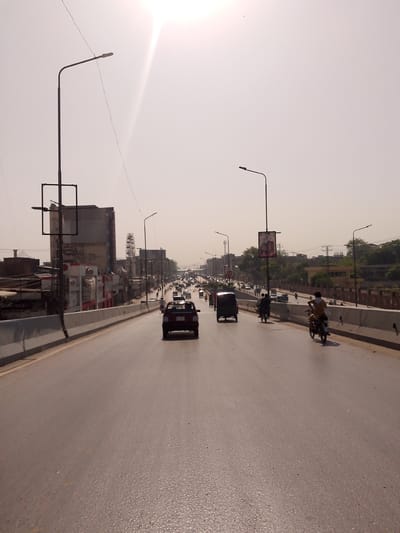 This is a wide-angle, eye-level shot taken on an elevated roadway in Peshawar City Tehsil, Pakistan. The road is multi-lane and paved with asphalt, reflecting the bright, hazy sky. Traffic is visible, including several cars and motorcycles. On the left side of the road, there is a concrete barrier and beyond that, buildings and a streetlamp. On the right side, there is also a concrete barrier, and farther beyond, buildings and trees are visible. A person on a motorcycle is in the foreground on the right, moving away from the camera. A lone motorcycle is in the middle ground, also moving away. Several cars are further down the road. The sky is predominantly white and bright, suggesting it is daytime, possibly midday or late morning. The overall scene depicts a busy urban environment on a highway.