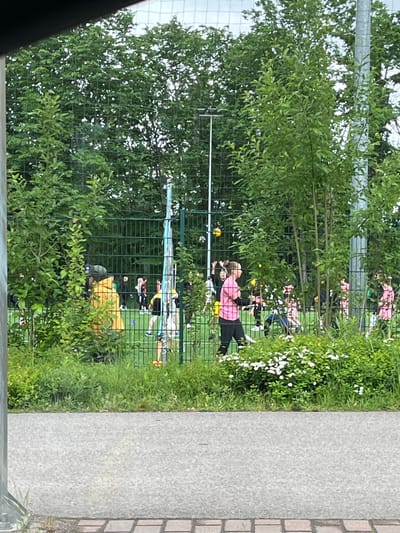The image depicts an outdoor sports field with a fence in the foreground. Several individuals, appearing to be primarily young women, are engaged in activities on the field. Some are wearing pink tops and dark bottoms. There are multiple volleyballs visible, suggesting a game or practice is underway. The environment includes green trees and bushes, with a paved area and a brick-like pathway in the foreground. The scene is consistent with a park or sports facility. No specific landmarks or identifying features indicate the city of Helsinki, Finland.