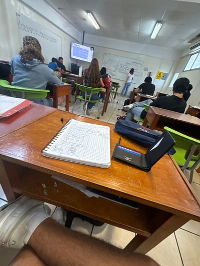 The image shows a classroom setting. There are students sitting at wooden desks, presumably listening to a lecture or lesson being presented on a screen at the front of the room. The classroom appears to be in use, with notebooks, pens, and other school supplies visible on the desks. The specific location is unknown, but based on the presence of Spanish in the text visible in the image it might be in Acapulco, Mexico. The image captures a snapshot of a typical learning environment.