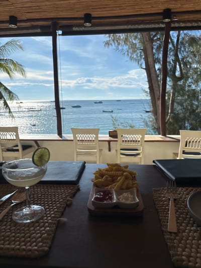 The image depicts a static scene inside an open-air restaurant or bar in Koh Tao, Thailand, providing a direct view of the ocean. In the immediate foreground, a dark wooden table is set with two woven placemats. On the left placemat, a stemmed cocktail glass, likely a margarita, holds a clear beverage garnished with a lime slice and features a salted rim. A fork, knife, and spoon are positioned next to it. On the right, a small wooden serving board presents a white ceramic dish divided into two sections, containing crinkle-cut french fries, a dollop of ketchup, and a dollop of mayonnaise. A silver fork and a grey plate are visible on the right placemat. Two dark-colored menus or binders rest on the table.
The overhead structure features a bamboo-slatted ceiling with two visible black cylindrical spotlights. A dark wooden support pillar is centrally located in the midground, framing the expansive sea view. Beyond the table, on a lower outdoor patio, several white, lattice-backed chairs are unoccupied, facing the water.
The background shows a vast expanse of blue ocean under a partly cloudy sky. Sunlight reflects brightly on the water's surface. Numerous boats, including smaller longtail-style vessels and larger motorboats, are visible scattered across the water. On the far left, a palm frond is partially visible, while the right side of the frame shows dense green foliage from trees bordering the coastline. The scene conveys a bright, clear day by the sea.