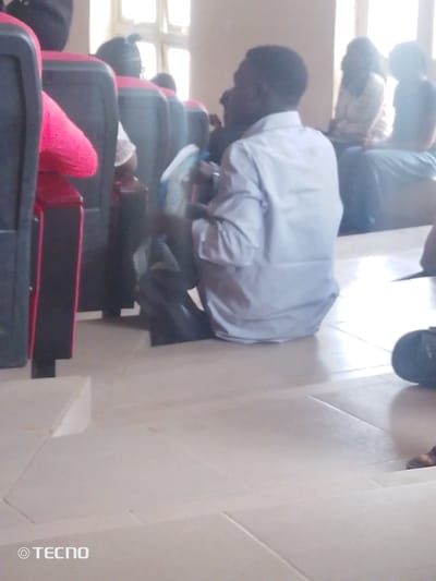 In a room with large windows casting bright light, a man in a light-colored collared shirt and dark trousers sits on the floor in the foreground. He is facing away from the camera and appears to be holding a blue and white object. Behind him, several other individuals are seated in rows of chairs, their faces and details obscured by the lighting and distance. The flooring is tiled in a light color. A black object is visible on the floor to the right of the man. The scene is likely within the city of Gumel, Nigeria, possibly in a lecture hall or meeting room due to the seating arrangement.