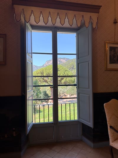 The image shows an open window in what appears to be an interior setting in Bunyola, Spain. The window is divided into multiple panes, offering a view of a landscape with mountains, lush greenery, and fields. The sky is clear and blue. The interior has patterned wallpaper, and a painting hangs above the window on one side. The window frame is white, and the walls are decorated with a unique style of trim. Parts of a chair are also visible next to the window.