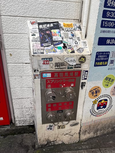 The image depicts a street scene in Shinjuku, Japan, focusing on a metal fire hydrant unit attached to a building wall. The hydrant is covered in various stickers, predominantly anime-themed and artistic decals. Prominently displayed is a red sign with Japanese text, "高田馬場駅" (Takadanobaba Station), indicating the location. The hydrant has two silver metal caps with chains. The wall to the right of the hydrant features a blue sign with white Japanese text indicating clinic hours and services, including "受付" (Reception) and "休診" (Closed). Below the blue sign is a circular sticker with a smiling cartoon train and the text "TAKADANOBABA E.N.T. CLINIC". The ground in front of the hydrant is a cracked concrete sidewalk with some moss and debris. To the left of the hydrant, a portion of a red metal structure, possibly a mailbox or a door, is visible. The overall impression is an urban environment with elements of local culture and signage.