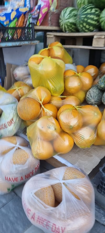 In Şïeli, Kazakhstan, a street vendor is seen selling a variety of fruits. The scene depicts an outdoor market stall laden with watermelons, oranges, and melons. The watermelons, in shades of green, are stacked on wooden pallets. Oranges are packaged in bright yellow plastic bags, with some also in clear plastic. Melons, with their characteristic netted rind, are also bagged in clear plastic, with some bags bearing the text "HAVE A NICE DAY" in red lettering. A black crate is visible in the background, containing more produce. The ground is paved with grey stones, suggesting a public walkway or market area. The overall impression is one of a bustling local market, offering fresh produce to passersby.