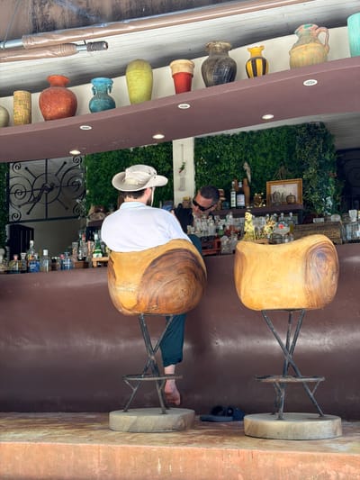 The image depicts a man wearing a white hat and white shirt sitting on a stool in what appears to be an outdoor establishment, possibly a bar or restaurant, in Baan Thong Krut, Thailand. The man is facing away from the camera, his body angled to the right. He is seated on a large, carved wooden stool with a metal base. In the background, shelves are filled with various bottles, suggesting a bar setting. There is also a lush green wall of foliage, a common decorative element in tropical locations. The overall scene suggests a relaxed atmosphere, potentially during the daytime given the brightness. The man's posture and attire are casual. The image is taken from a slightly elevated perspective, looking down and to the side. The overall color palette is warm, with earthy tones from the wood and building materials contrasted by the vibrant green of the plants and the varied colors of the bottles.