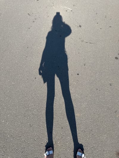 A full-body shadow of a person is cast on an asphalt surface. The shadow is elongated, indicating a low sun angle. The person appears to be standing, wearing shorts and a top. A bag or purse is visible hanging from their left side. The asphalt is grey and textured, with scattered dark spots that could be stains or debris. At the bottom of the image, the shadow's feet are visible, wearing white sandals with dark soles and silver buckles. The scene is likely outdoors on a sunny day, possibly a sidewalk or street in Cheboksary, Russia, given the reference to the location.