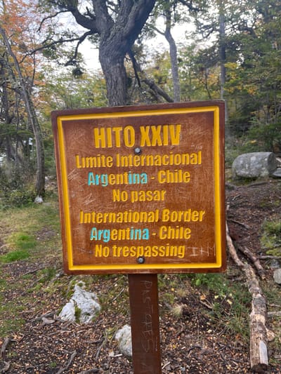The image shows a weathered, brown and yellow sign marking the international border between Argentina and Chile. The sign, titled "HITO XXIV," displays text in both Spanish and English, indicating "Limite Internacional Argentina - Chile" and "International Border Argentina - Chile," along with "No pasar" and "No trespassing." The sign is mounted on a brown wooden post, and the word "PASTORES" is faintly scratched into the post below. The sign is situated in a natural environment with trees and ground covered in leaf litter and vegetation. The surrounding environment suggests a forested area, possibly a park or hiking trail. The image is taken at ground level, and the perspective is slightly tilted. The location is Lapataia, Argentina.