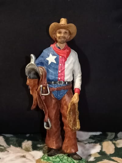 This is a medium-full shot of a decorative figurine of a cowboy. The figurine stands upright on a base that appears to be made of resin or painted plaster, depicting rocky terrain with some patches of green and white. The cowboy is wearing a brown cowboy hat, a red bandana, and a shirt that is split vertically down the middle. The left side of the shirt is blue with a white star, resembling the Texas flag, while the right side is white with red buttons. He also wears brown chaps over blue pants and brown boots. He is holding a coiled rope in his right hand and a saddle and bridle in his left. The background is a solid black fabric. The figurine is well-lit and in focus. There is no information to suggest this image is located in Amarillo, United States of America.