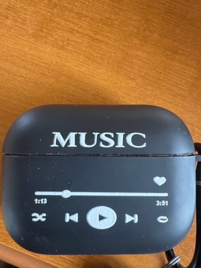 The scene is a close-up, top-down view of a black, matte silicone protective case for wireless earbuds, resting on a wooden surface in Fowlis Easter, United Kingdom. The case, exhibiting minor dust particles and scuffs, features white embossed graphics. Prominently centered on the upper half, the word "MUSIC" is displayed in a serif font. Below this, a stylized music player interface is depicted: a horizontal progress bar shows a scrubber positioned at "1:13" out of "3:51," with a white heart icon above the right end of the bar. Beneath the progress bar, from left to right, are a shuffle icon, a previous track icon, a play button (triangle within a circle), a next track icon, and a repeat icon. A black lanyard or strap, secured by a small metal ring, is attached to an integrated loop on the right side of the case. The background is a warm, light brown wooden surface, likely a desk or table, with visible grain, rendered slightly out of focus towards the top of the image. No individuals, discernible actions, or interactions are present in the static composition.