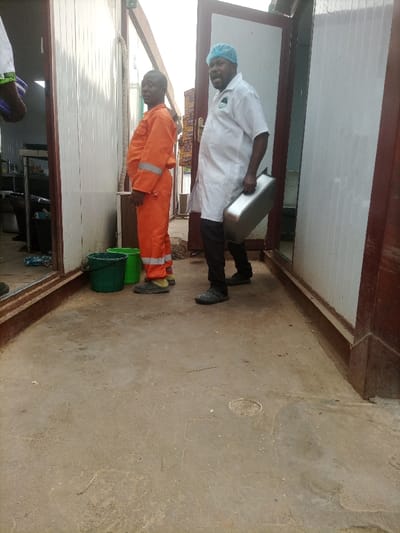 Two men stand in a narrow, enclosed space between buildings. One man, wearing an orange jumpsuit, stands near two green buckets. The other man, wearing a white lab coat, black pants, and a blue hair net, carries a silver container. Both men wear dark footwear. The concrete floor is stained. The buildings have white paneled walls with brown trim. One building appears to house a kitchen. A partially opened door provides a glimpse to the outdoors.