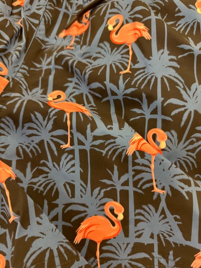 The image is a close-up of a fabric with a tropical pattern. The pattern consists of stylized orange flamingos and silhouettes of palm trees against a dark background with blue palm fronds. The fabric appears to be part of a garment, possibly swimwear, as suggested by the drawstring visible at the top left. There are no people or specific locations within Steenwijk, Netherlands, identifiable in this image.