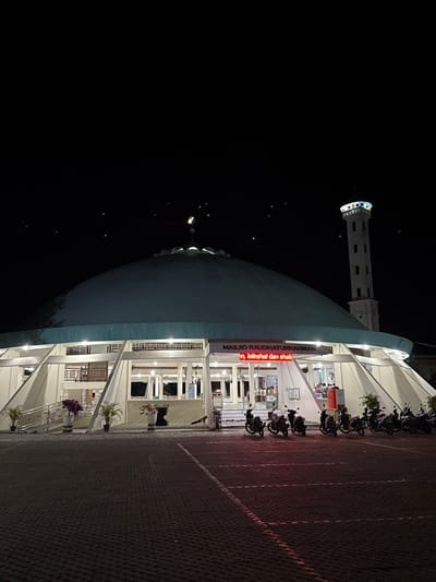 Here's a description of the image:
The image is taken at night of the Masjid Raudhaturrahmah, a mosque in Pasar Paloh, Indonesia. The mosque has a large dome and a tall minaret. A brightly lit LED sign with red lettering is visible on the mosque's facade. Motorbikes are parked in a row along the mosque's edge on a brick paved surface. The sky is black.