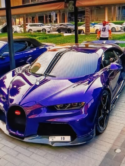 A purple Bugatti Chiron is parked on a tiled surface in front of a modern building. The car has a black front grille with a red Bugatti emblem and a license plate that reads "DUBAI 10". A man in a white t-shirt with a graphic print is standing in the background, looking at the car. Other cars, including a white SUV and a yellow sedan, are parked in the background. The environment appears to be an outdoor parking lot or showroom area, possibly in Dubai. The image does not contain information about the city of Sluck, Belarus.