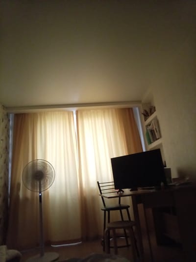 The image depicts an indoor scene, likely a living room or bedroom, in Cheboksary, Russia. The room is dimly lit, suggesting it could be evening or a cloudy day. The dominant features are a large standing fan on the left, positioned in front of sheer, light-colored curtains that cover a window. The curtains are illuminated from behind, creating a soft glow.
To the right of the fan, a small table is set up with a chair and a stool. On the table sits a dark computer monitor, facing away from the viewer. A shelf is mounted on the wall above the table, displaying several items, including books and possibly decorative objects.
The overall impression is of a functional space, perhaps a home office or a cozy corner. The lighting conditions and the presence of a fan hint at warm weather. There is no visible text in the image. The scene is quiet and still, with no people or overt activity taking place. The environment appears somewhat cluttered with items on the shelf and the general arrangement of furniture.