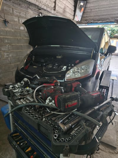 A black Citroen C4 Picasso sits with its hood open in a garage in Moaña, Spain. The vehicle is positioned on a mechanical lift. In the foreground, a toolbox laden with an assortment of tools, including sockets, wrenches, and power tools, occupies the space in front of the car. The garage interior features cinder block walls. No people are visible. The focus of the scene is on car repair and maintenance.