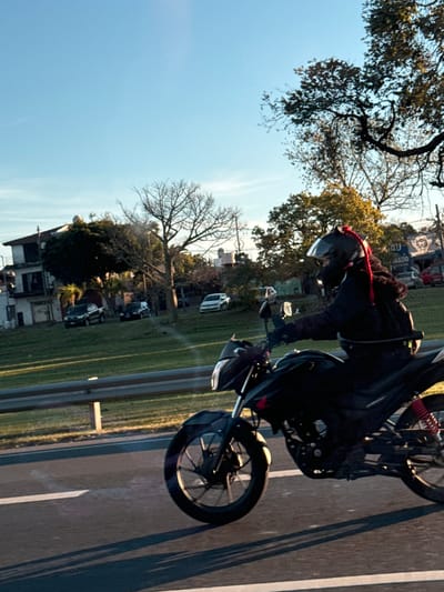 The image depicts a motorcycle with a rider in Virreyes, Argentina. The rider wears a black suit and helmet, accented with a red braided cord. The motorcycle is traveling on a paved road with white lane markings, adjacent to a grassy area. Buildings and bare trees are visible in the background, set against a blue sky. Several vehicles are parked near a house, and a larger truck with signage on its side is also present. The overall environment suggests an urban or suburban street scene during daylight.