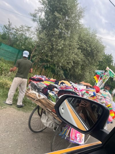 In Gulab Bagh, India, a man wearing a light-colored cap and a dark green t-shirt stands on a dirt road, facing away from the camera. He is wearing light-colored trousers. Beside him is a cart overflowing with various items, including folded clothes, bags of merchandise, and cleaning supplies such as brooms and brushes. A second man, wearing a light-colored shirt, is visible behind the cart, looking towards the camera. The background consists of lush green trees and a cloudy sky. The scene is captured from inside a vehicle, as indicated by the reflection of the car's interior and occupants in the side mirror.