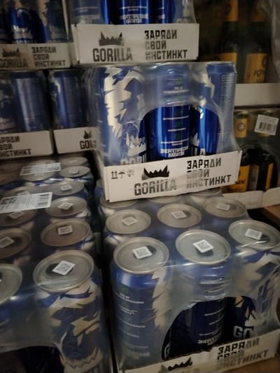 This indoor photograph captures multiple stacked cases of "Gorilla" brand energy drinks, likely in a warehouse, storage room, or retail backroom in Заволжье, Russia. The scene primarily consists of these product packages, with no people or specific activity taking place.
Each case holds several blue-colored cans, packaged together with clear plastic wrap on a cardboard tray. The cans feature white and lighter blue graphical patterns. The brand name "GORILLA" is prominently displayed on the cardboard trays, accompanied by a stylized crown logo. Below the brand name, the Cyrillic slogan "ЗАРЯДИ СВОЙ ИНСТИНКТ" (CHARGE YOUR INSTINCT) is clearly visible on multiple trays. Some cans also show text like "ЭНЕРГЕТИЧЕСКИЙ НАПИТОК" (ENERGY DRINK), and smaller details such as "СОДЕРЖИТ ВИТАМИНЫ" (CONTAINS VITAMINS) and "ЭКСТРАКТ ГУАРАНЫ" (GUARANA EXTRACT) can be discerned on the can labels. Small QR codes are affixed to the tops of many cans. One cardboard side displays shipping symbols for "fragile" and "keep dry."
In the background, out-of-focus shelves hold other items, including bottles with yellow labels, suggesting a variety of beverages or products are stored in the same area. The lighting appears to be artificial, casting some shadows and giving the scene an industrial or commercial storage feel. The overall color palette is dominated by the blues of the cans, the brown of the cardboard, and the grey/silver of the can tops.