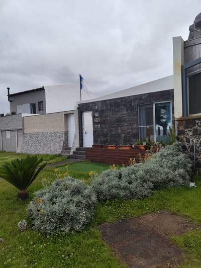The image depicts a cloudy day in Ponta Delgada, Portugal, with a view of a modern house. The house features a dark, textured facade and large windows, with a white section and a gray upper level. In front of the house, there is a garden with various plants, including a palm-like cycad, grey-green bushes, and small yellow flowers. Several potted cacti are displayed on a wooden planter. A flag with blue and white sections, and a yellow cross, flies from a pole on the roof. The sky is overcast, suggesting a potentially damp or cool day. The scene is peaceful and suburban, with no people visible. There is some text on a wooden sign hanging on the right side of the house, which appears to be in Portuguese.