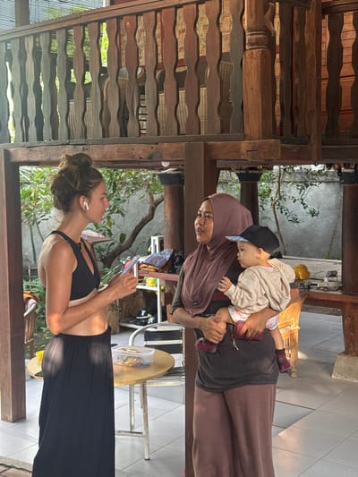 In Pemenang Timur, Indonesia, two women and a baby are interacting outdoors. The woman on the left, wearing a black sports bra and a long black skirt, holds a small device in her hands. She has her hair pulled up in a bun and is wearing white wireless earbuds. The woman on the right, dressed in a brown hijab and a brown long-sleeved shirt with a darker panel, is holding a baby. The baby, wearing a light-colored hooded jacket, a patterned diaper cover, and socks, is looking to the right. They are standing under a wooden structure, possibly a verandah or an elevated platform with ornate wooden railings. In the background, there are trees, a gray wall, and furniture, including chairs and a round table with a clear container filled with food. The scene appears to be in a residential or communal outdoor area.