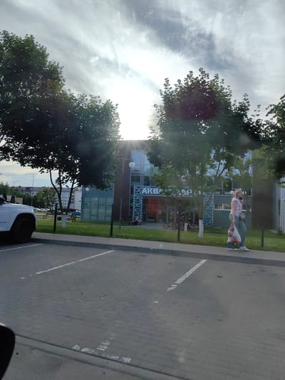 The image shows a bright, overcast day in Fanipal, Belarus. The scene is a parking lot in front of a modern building with a glass facade. The building's exterior has large windows and a dark frame. A prominent blue sign with white lettering on the building reads "AKBA".
In the foreground, a white SUV is parked on the left side of the frame. The parking lot is paved with interlocking bricks. White lines mark the parking spaces. The ground is clean and dry, with a few scattered leaves.
To the right of the building, a woman with blonde hair is walking along the sidewalk. She is wearing a light pink t-shirt and dark pants. She is carrying a white plastic bag with red lettering, and a light-colored purse or bag slung over her shoulder. She appears to be looking at her phone, which she holds to her ear. Her expression is neutral.
Behind the woman and the building, there are trees with lush green foliage, suggesting it is summer or late spring. The sky is filled with wispy clouds, with the sun partially obscured, creating a bright, diffused light. In the distance, behind a fence and some grass, several apartment buildings are visible, as well as parked cars. The overall impression is of an ordinary day in a suburban or commercial area.