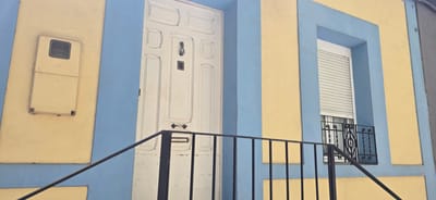 This is a medium shot of the exterior of a building with a distinctive color scheme of yellow and blue. The building features a white door with multiple panels, a door knocker, a mail slot, and a doorbell or intercom. To the left of the door, a cream-colored utility box is mounted on the yellow wall, which is bordered by blue. To the right of the door, a window is visible, covered by a white roller shutter. Below the window, a decorative black iron railing is present. A black metal railing also runs diagonally across the foreground, leading up to the entrance. The architectural style and the colors suggest a Mediterranean or Southern European setting, consistent with the provided location context of Huesca, Spain. There is no visible text in the image. The lighting suggests it is daytime, and the weather appears clear. The scene captures a humble, residential entrance.
