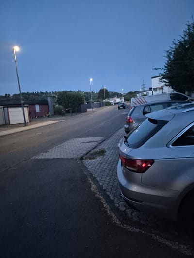 The image captures a street scene in Trier, Germany, during twilight. On the right, the rear of a silver station wagon is prominently featured, parked along a cobblestone sidewalk adjacent to an asphalt road. To its left, another silver station wagon is partially visible, followed by a dark-colored hatchback and a white van with red stripes. The road stretches into the distance, lined with parked vehicles and bordered by a sidewalk. On the left side of the road, a row of garages with light-colored doors stands beside a paved area. Further in the background, trees and low-rise buildings are visible against a darkening sky, illuminated by streetlights. The overall atmosphere is quiet and residential, with no people present in the frame.
