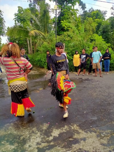 The image shows a group of people standing on a paved road in Kecamatan Kedawung, Indonesia (latitude: -7.514, longitude: 111.03).  There are two people in the foreground. The person on the left is wearing a red and white striped shirt with a yellow and black skirt. They have a black strap around their waist. The person on the right is wearing a black shirt and black pants. They have a brightly coloured mask made of red, yellow, and green.  There are other people in the background,  wearing various attire.  The image is taken from a slightly elevated angle. The road is wet, and there are pink flower petals scattered on it.  The background includes lush green trees and a light pole.