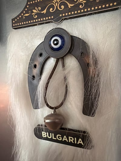An overhead shot captures an arrangement of decorative items resting on a white, fluffy, fur-like surface, potentially in Lusail, Qatar. The central object is a dark, metallic or wooden horseshoe, featuring several small circular holes along its edges. At the top center of the horseshoe, an "evil eye" charm is attached, depicted with concentric circles of blue, white, and a black pupil. A thin brown leather cord is threaded through the horseshoe, looping around and ending with a small, shiny, tear-drop shaped bead or charm, possibly metallic.
To the right of the horseshoe lies a slender, rectangular, dark-colored tag oriented vertically, displaying the word "BULGARIA" in prominent gold or light-colored capital letters. To the left, partially visible, is a section of another dark, flat object, likely wood, adorned with intricate gold-colored floral or scroll patterns and a border of small, evenly spaced dots. The overall scene is a still life, showcasing these cultural and decorative pieces on a soft, neutral background. No people are visible, and no specific time of day or weather is indicated.