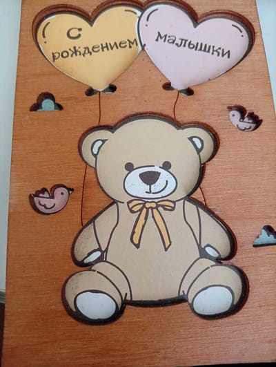 This is a close-up, eye-level, medium shot of a wooden plaque with a decorative design. The plaque is made of light brown wood with a visible grain. The central subject is a cartoonish teddy bear, depicted from the waist up, sitting with its legs extended. The bear is light brown with black outlines and has a yellow ribbon tied around its neck. Its face is visible, featuring a black nose and smiling mouth, and white inner ears and cheeks.
Above the bear are two heart-shaped balloons, also with black outlines. The left balloon is yellow and inscribed with the Russian text "С рождением" (meaning "Congratulations on the birth"). The right balloon is pink and inscribed with "Малышки" (meaning "Little ones"). To the left of the yellow balloon and above the bear's left arm, there is a small, light blue cloud-like cutout. To the right of the pink balloon and above the bear's right arm, a small, light pink bird cutout is visible. Another similar bird cutout is located to the left of the bear's left arm, and a light blue cloud cutout is above the bear's right leg.
The image does not contain any information that allows for its location in the city of Agoy, Russia.
