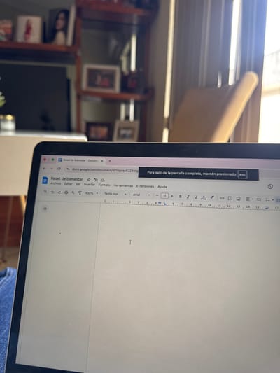 The image displays an indoor scene located in Ciudad Victoria, Mexico, with a laptop computer occupying the lower half of the frame. The laptop screen shows a Google Docs interface open to a blank document titled "Reset de bienestar," with a blinking cursor present on the page. A dark banner at the top right of the screen instructs the user, in Spanish, to "hold down esc" to exit full-screen mode. A portion of a person's blue denim-clad lap or leg is visible in the bottom left, suggesting the laptop is situated on the user's lap. The background is blurred due to shallow depth of field. To the left, a dark television screen is visible beneath a multi-tiered wooden shelving unit. The shelves contain various decorative items, including multiple framed photographs, one featuring a woman, and another appearing to be a family portrait. To the right of the laptop, a bright window or glass door allows strong natural light into the room, illuminating a light-colored chair and partially revealing an exterior railing structure.